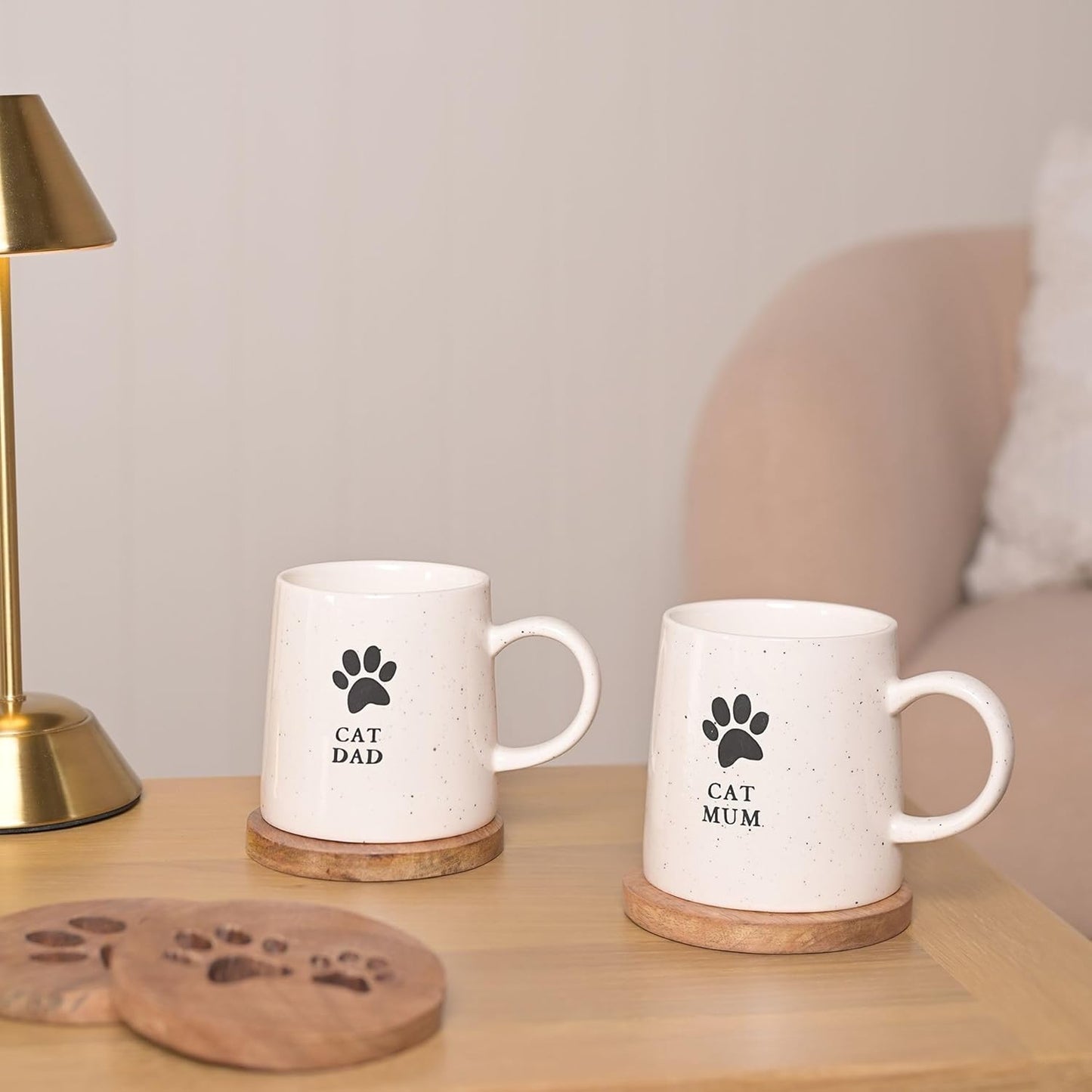Two white mugs with paw prints and text on a wooden surface next to a lamp.