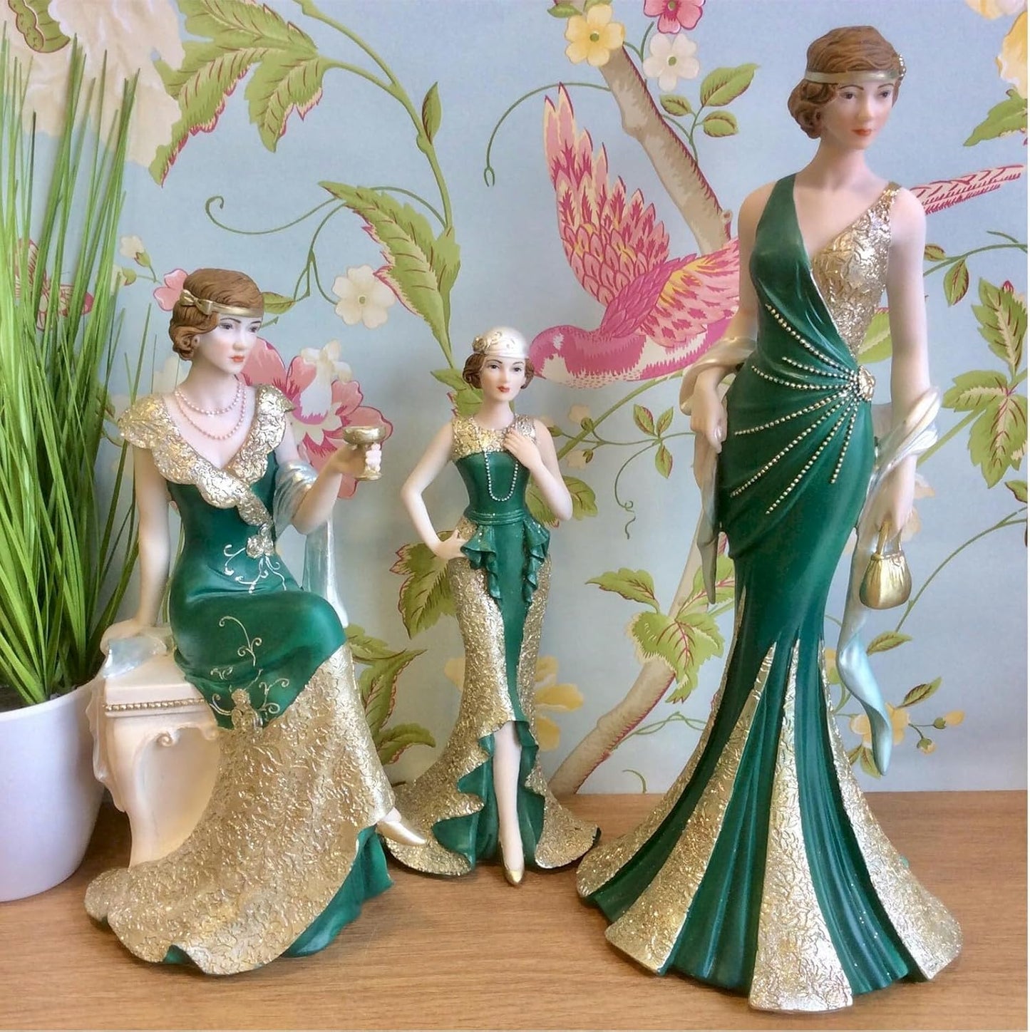 Three decorative figurines of women in elegant green and gold dresses against a floral background.