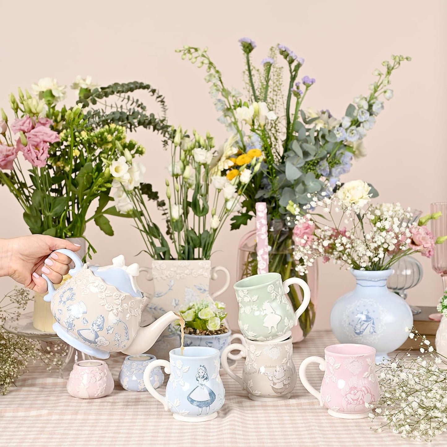 Pastel-colored ceramic tea set with mugs and a teapot on a table with floral arrangements.
