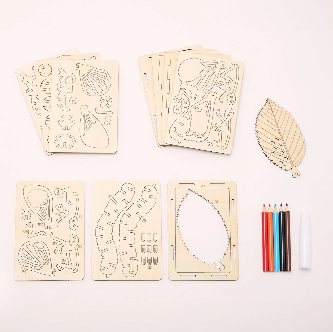 A leaf-shaped wooden DIY craft kit laid out on a white background, including multiple engraved wooden template pieces, a large leaf-shaped outline piece, colored pencils, and a paintbrush.