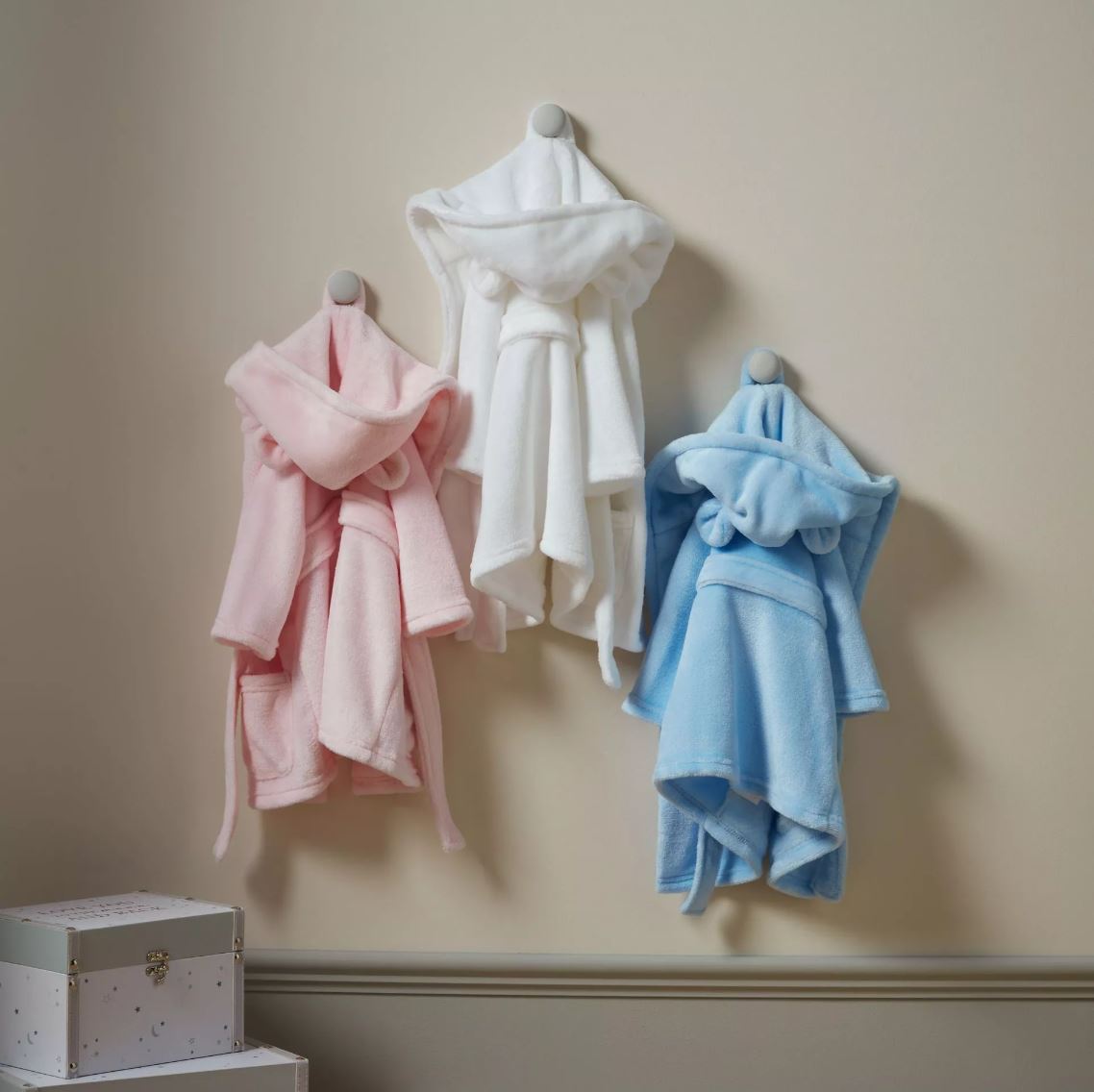 3 baby dressing gowns displayed on hooks in a house, pink white and blue