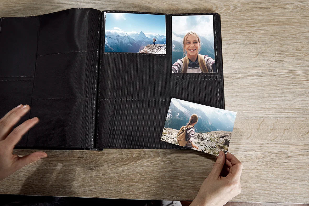 A black photo album binder open on a light wood table, with two sample landscape and portrait photos inserted and a third photo being placed into the album by hands.
