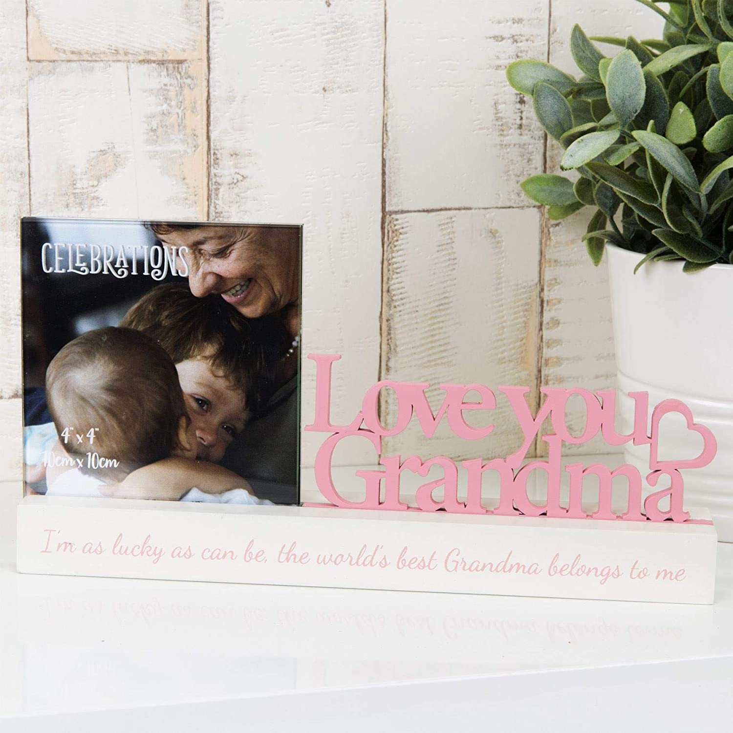 A pink 4x4 inch photo frame with 'Love You Grandma' inscription and a graphic of a grandmother hugging her grandchildren. In front is a photo of a grandmother with her grandchildren.