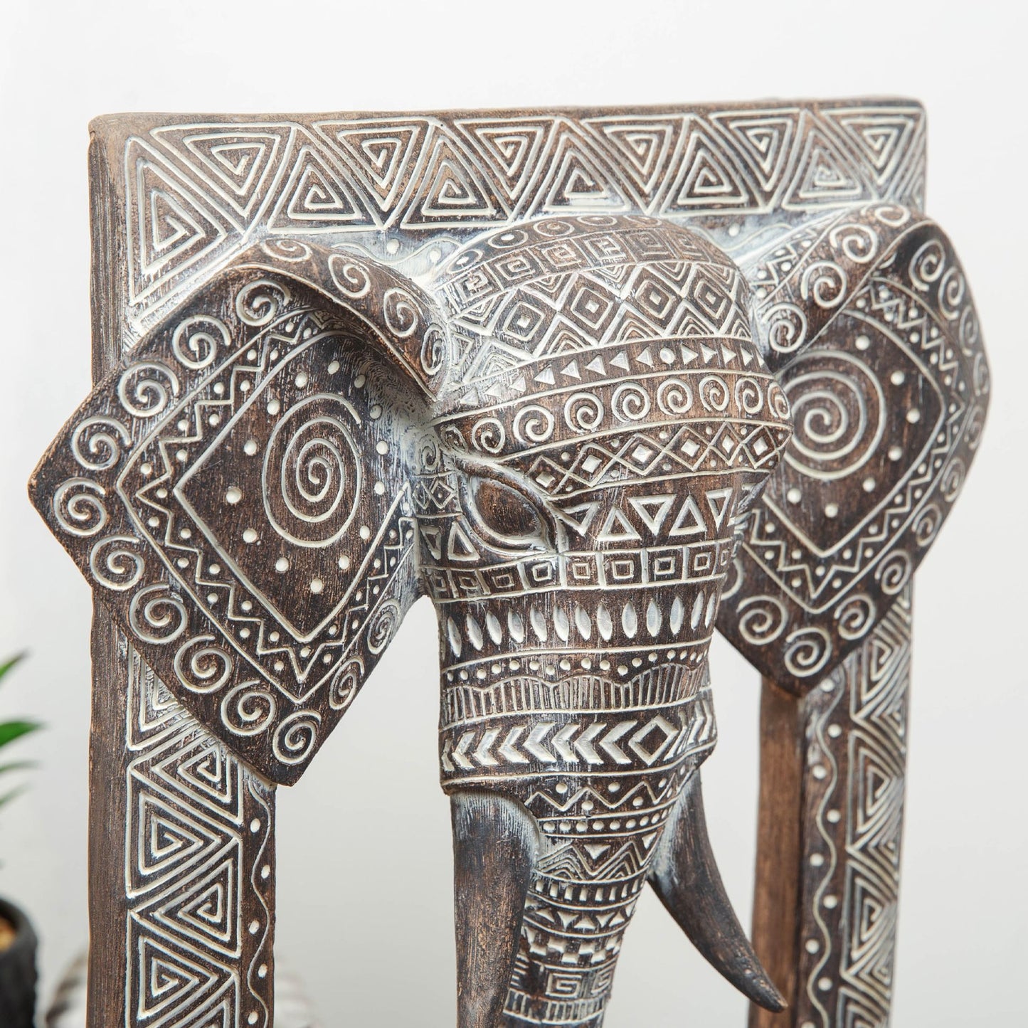 A brown, handpainted elephant sculpture with intricate carved patterns, standing on a base. close up
