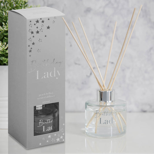 A Peach Bellini scented reed diffuser set, including a clear glass jar with a chrome finish metal lid and silver foil decal, accompanied by a box with 'Birthday Lady' text and star patterns.