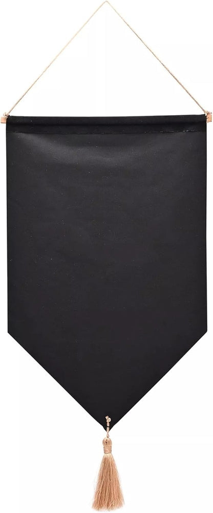 A black fabric wall hanging banner with a pointed bottom and a beige tassel, suspended by a thin beige cord with gold-toned metal rings, shown against a plain white background.