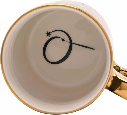 Close-up of the inside of a white ceramic mug with a gold rim, featuring a black monogram-style logo with stars and a wand printed on the cup, and a gold handle visible on the right.