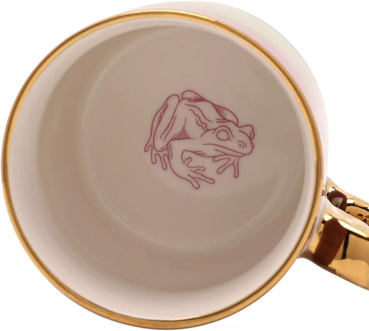 Close-up of the inside of a white ceramic mug with a gold rim, featuring a pink line-art illustration of a frog on the bottom and a gold handle.