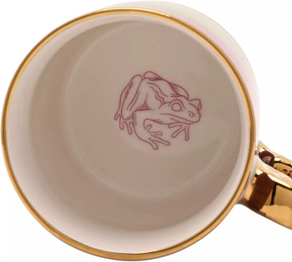 Close-up of the inside of a white ceramic mug with a gold rim, featuring a pink line-art illustration of a frog on the bottom and a gold handle.