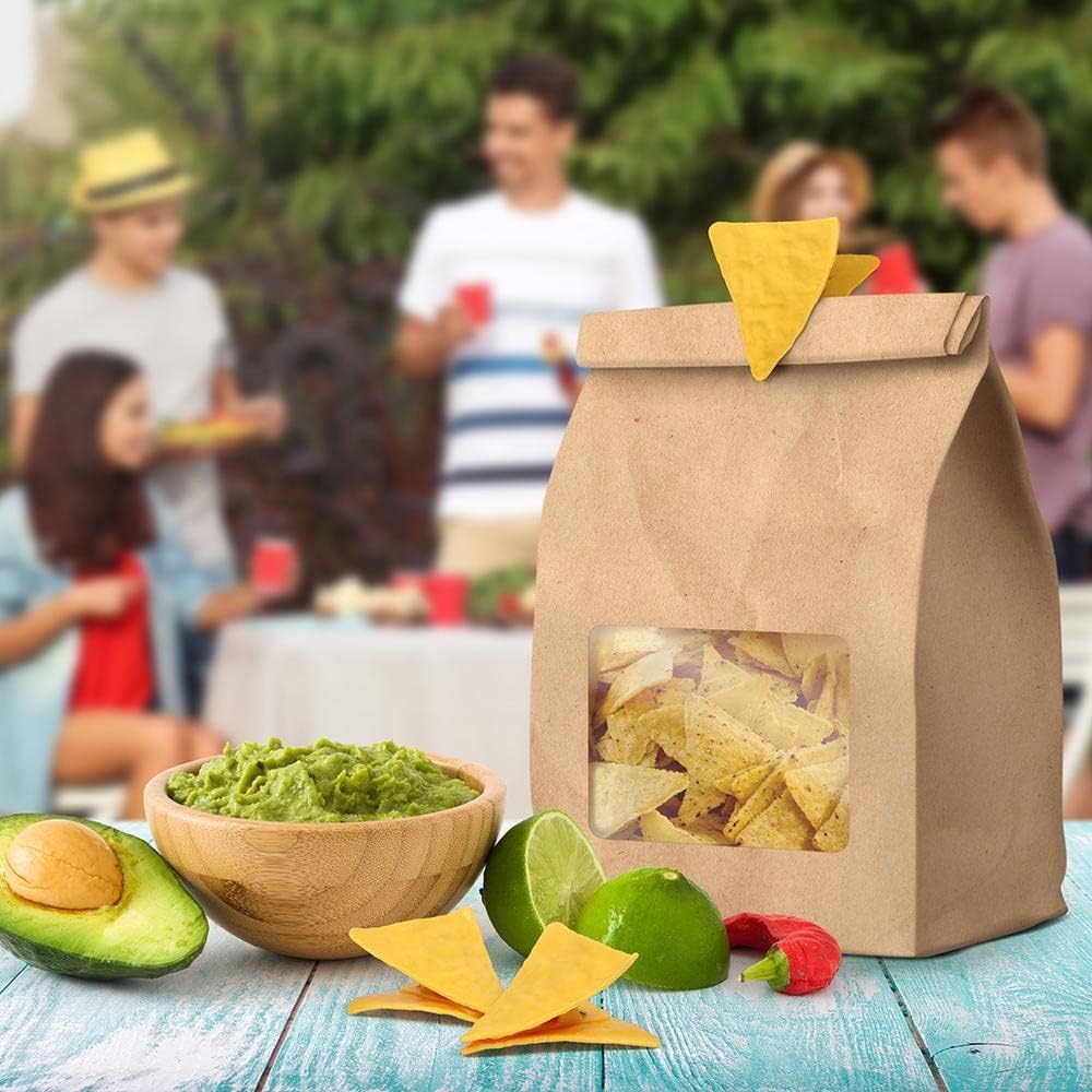 Kraft paper bag of tortilla chips with guacamole, avocados, and limes on a table with people in the background.