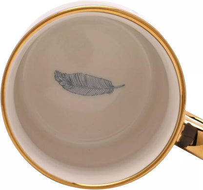 Top-down view of a white ceramic mug with a gold rim, a gold handle, and a blue feather illustration printed in the center of the inside.