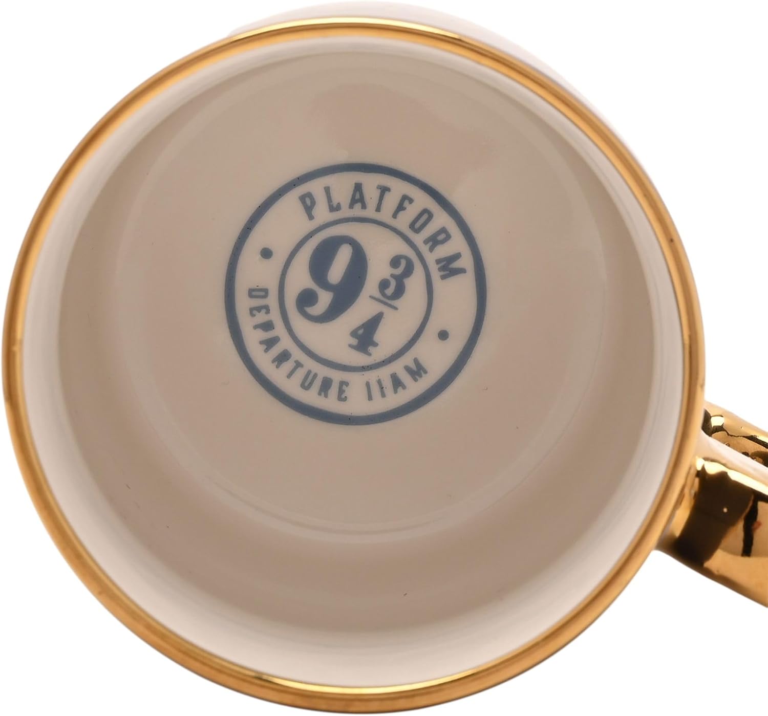 Close-up of the inside of a ceramic mug showing a printed blue circular stamp that reads 'PLATFORM 9 3/4 DEPARTURE 11AM' on a warm off-white background, with a gold rim and a gold handle visible on the right.