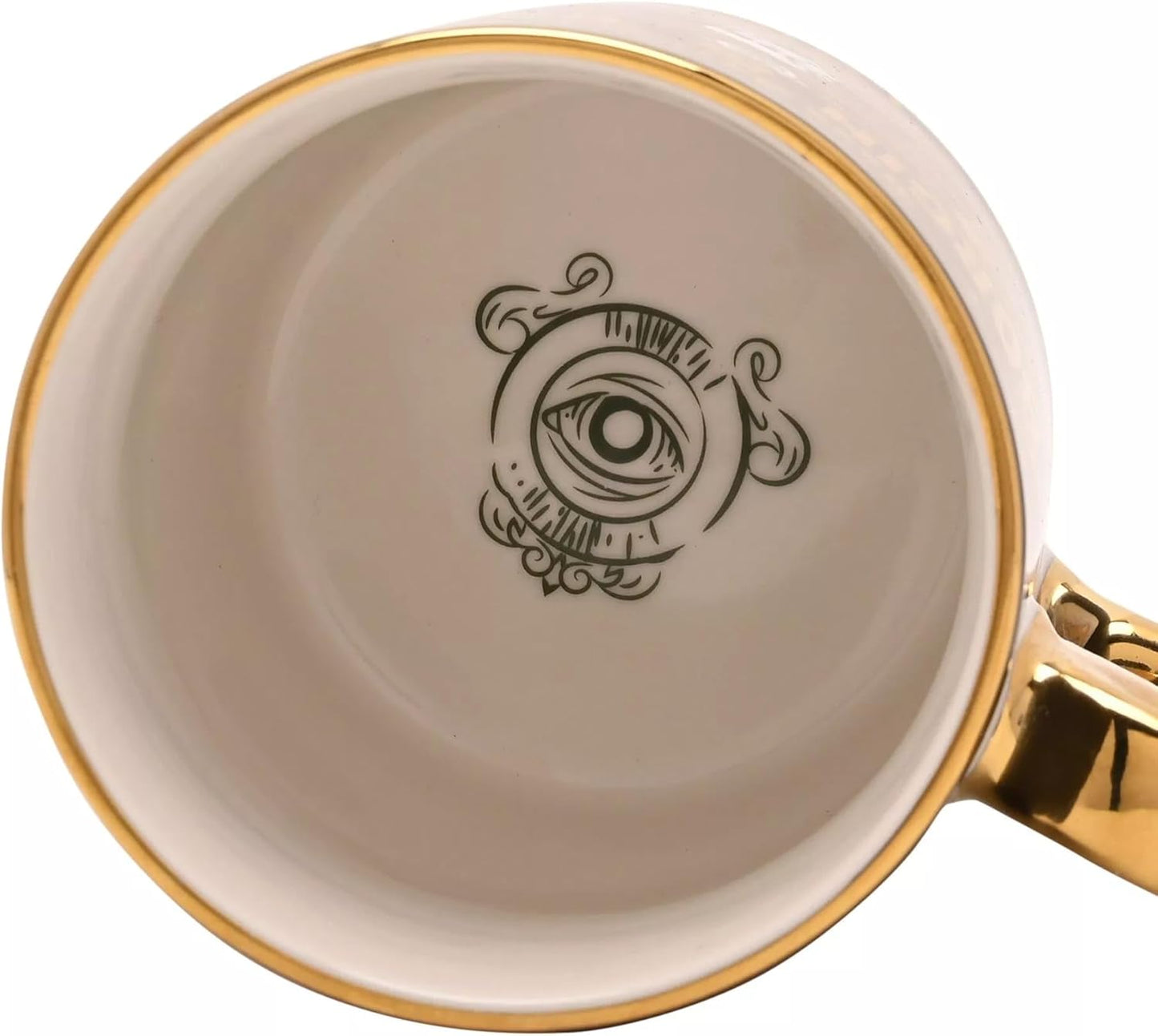 Close-up of the inside of a white ceramic mug with a gold rim, a gold handle, and a black eye illustration printed on the bottom.