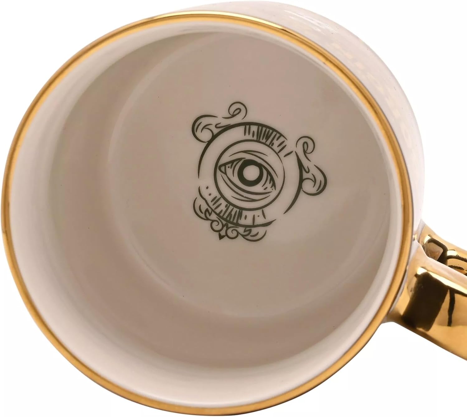 Close-up of the inside of a white ceramic mug with a gold rim, a gold handle, and a black eye illustration printed on the bottom.