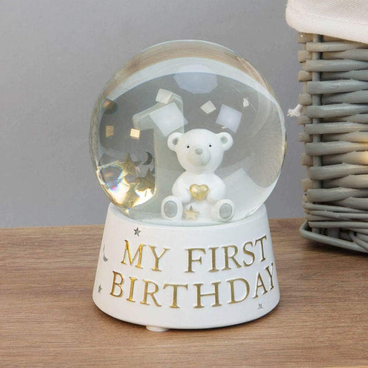 Snow globe with a teddy bear inside, labelled 'My First Birthday', on a wooden surface.