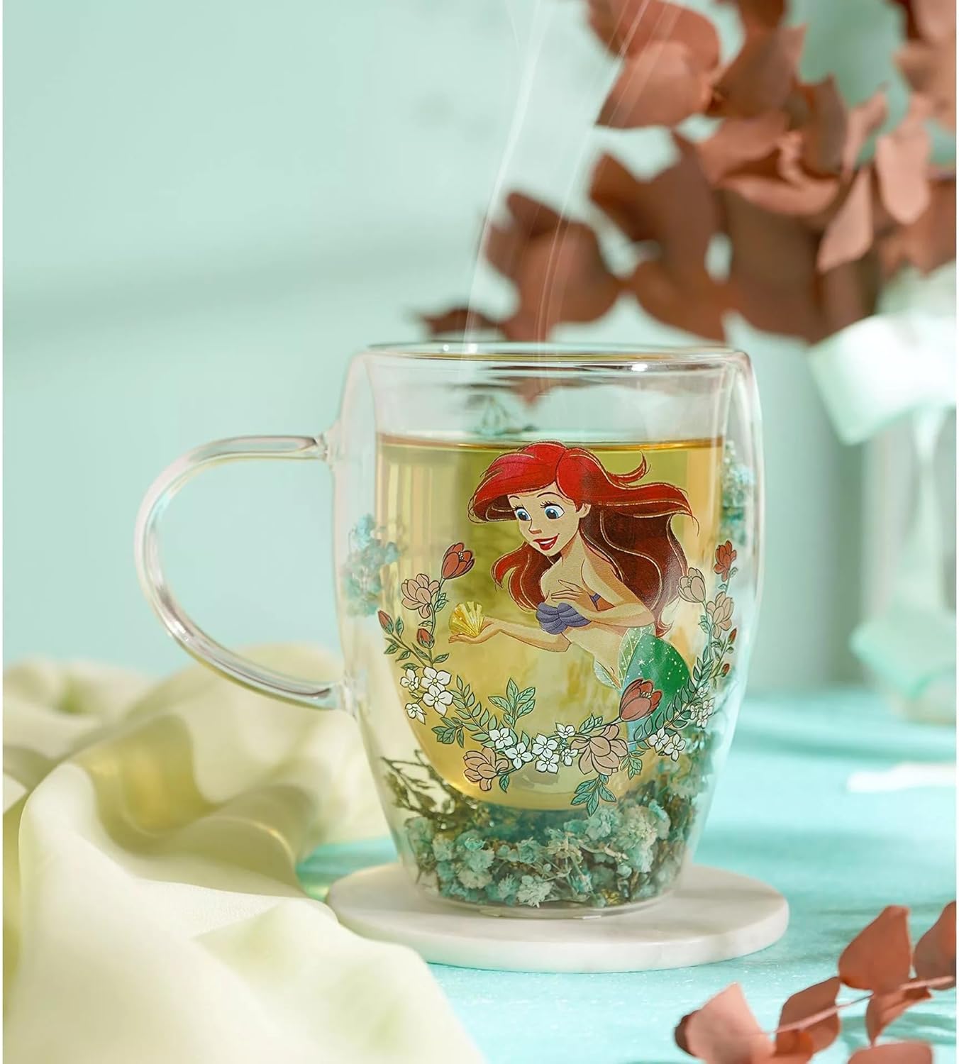 A clear glass double-walled mug with a handle, featuring a printed illustration of Ariel from The Little Mermaid on the front, filled with tea and placed on a white coaster. The background is a soft, dreamy pastel setting with light blue tones and decorative leaves.