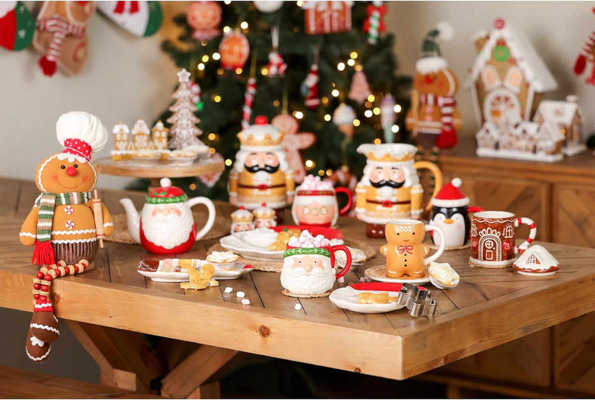 A collection of festive Christmas ceramic mugs, teapots, and gingerbread-themed table decorations arranged on a wooden table in front of a decorated Christmas tree.