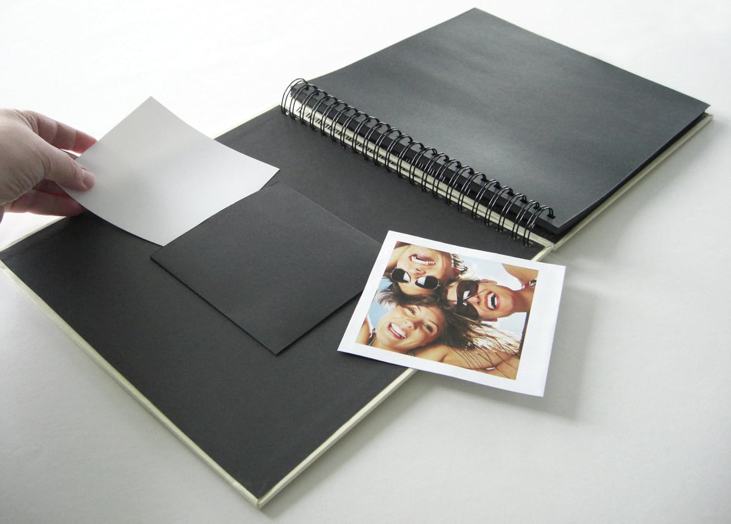 Black spiral-bound photo album with a hand holding a white card and a photo of two people on a white background.
