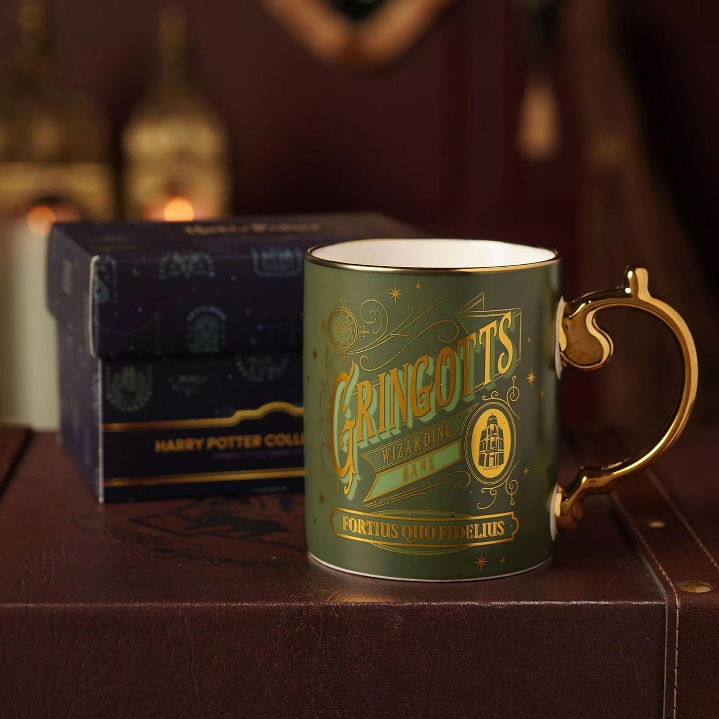A green ceramic Harry Potter Gringotts Wizarding Shop mug with a gold handle and gold lettering, displayed in front of its dark blue branded gift box on a brown leather surface.