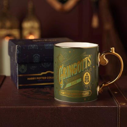 A green ceramic Harry Potter Gringotts Wizarding Shop mug with a gold handle and gold lettering, displayed in front of its dark blue branded gift box on a brown leather surface.