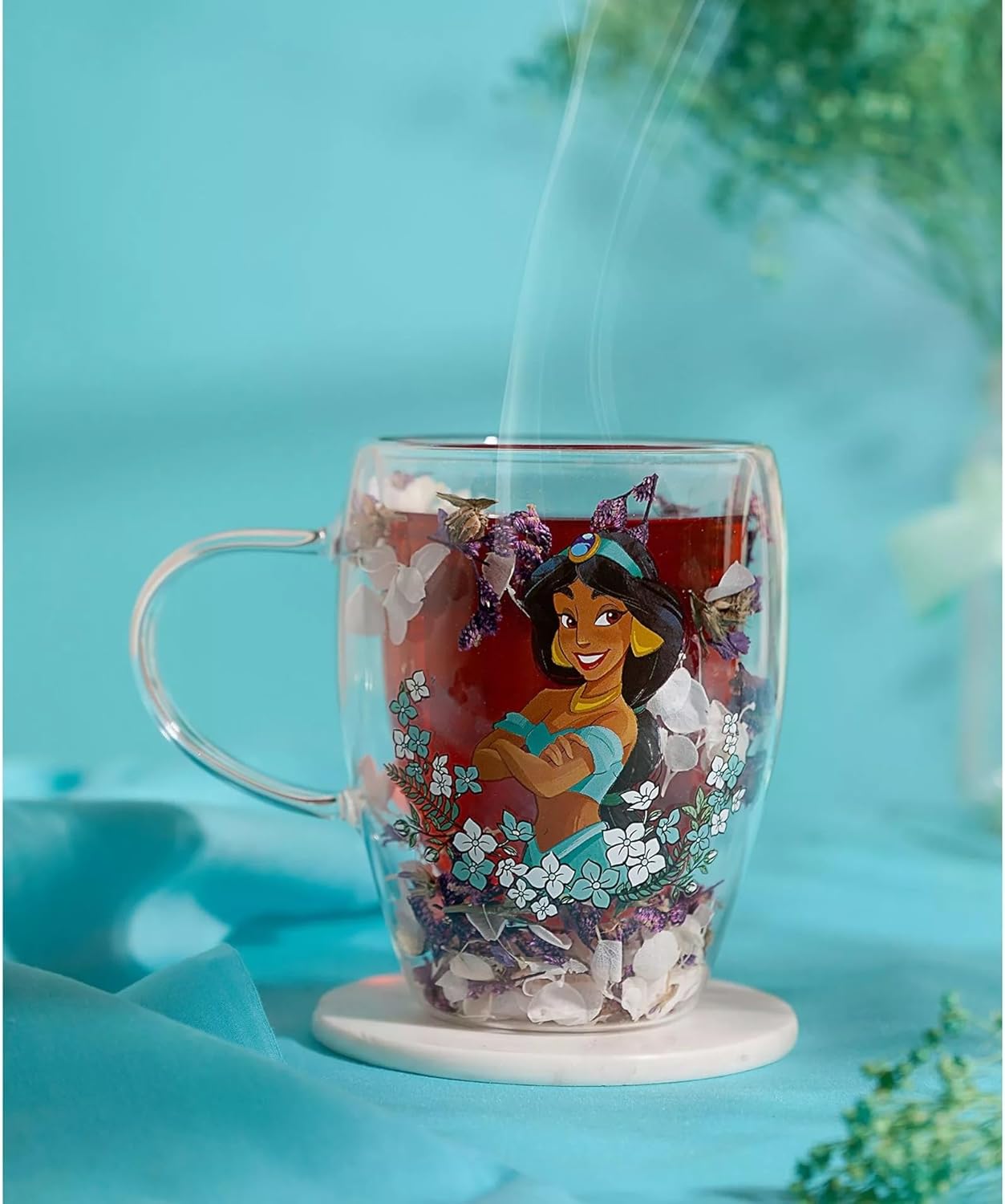 A clear double-walled glass mug with a white coaster base, featuring a printed illustration of Disney's Jasmine Al-Faruq. The mug is filled with red tea and decorative flower petals, with steam rising from the top, set against a soft turquoise blue background with blurred green foliage.