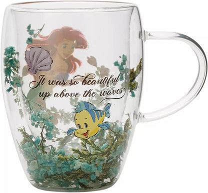 A clear double-walled glass mug featuring characters from The Little Mermaid, including Ariel, Sebastian, and a purple shell, with the quote 'It was so beautiful up above the waves' printed on the front.