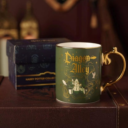 A green ceramic Harry Potter collectible mug with gold detailing reading 'Dragon Alley,' placed in front of a branded blue box on a dark brown surface.
