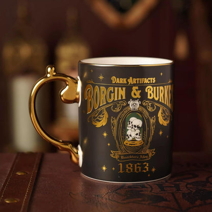 A dark ceramic mug with a gold handle and gold printed gothic-style artwork reading 'Dark Artifacts Borgin & Burles 1863' and 'Blackwood Alley,' shown on a brown leather surface with a blurred warm background.