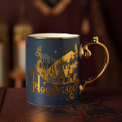 A blue ceramic mug with a gold metallic 'Hogsmade' castle graphic and a gold handle, set against a dark, warm, blurred background.