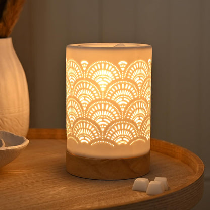 A cylindrical LED candle warmer with a warm glowing fan pattern cutout design, sitting on a round wooden tray next to a ceramic bowl.