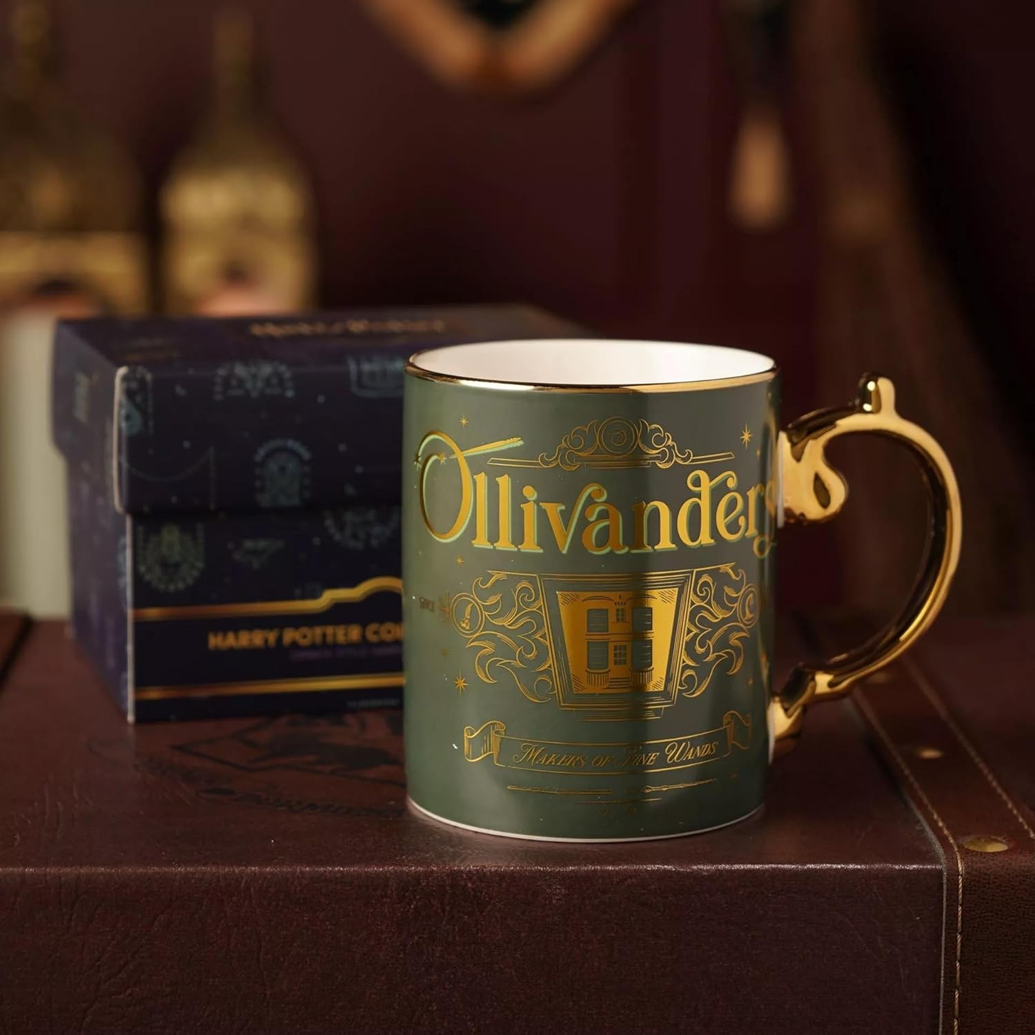 A dark green ceramic mug with gold foil detailing reading 'Ollivander' and a gold handle, shown with a dark blue Harry Potter Co-branded box in the background.