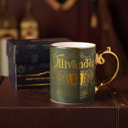 A dark green ceramic mug with gold foil detailing reading 'Ollivander' and a gold handle, shown with a dark blue Harry Potter Co-branded box in the background.
