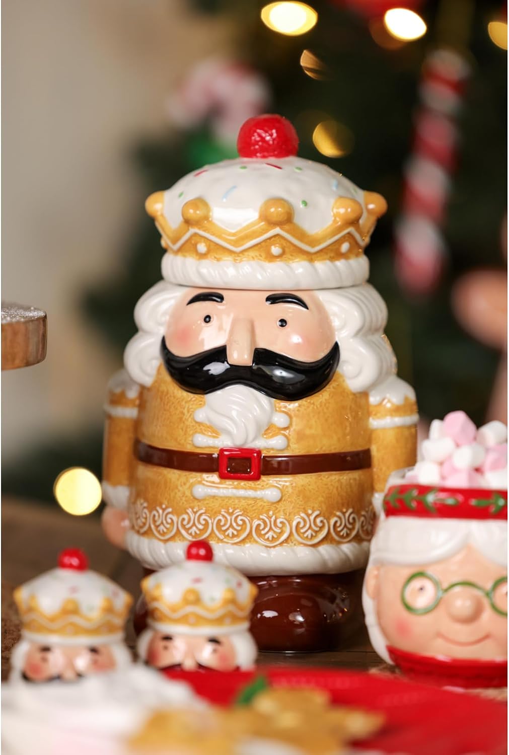 A ceramic Christmas nutcracker figurine with a gold and white outfit, black mustache, and red crown accent, shown in a festive holiday setting with blurred lights and other holiday decorations.