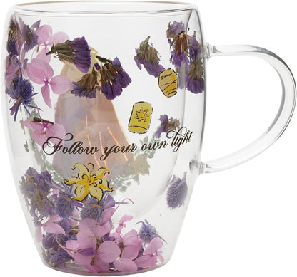 A clear double-walled glass mug with a handle, decorated with dried purple and pink flowers, yellow accents, and the printed phrase 'Follow your own light' in black script.