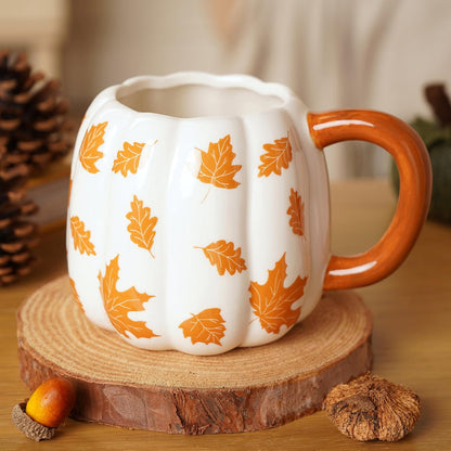 A white ceramic pumpkin-shaped mug with orange leaf patterns and a glossy orange handle, displayed on a rustic wood slice coaster with autumn-themed props.