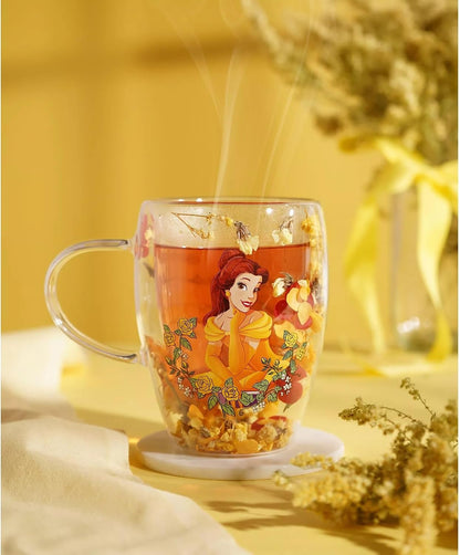A clear glass mug with a handle, featuring a colorful illustration of Belle from Beauty and the Beast on the front, filled with amber tea and dried flowers. The mug sits on a small white coaster on a yellow table with a warm, cozy background.