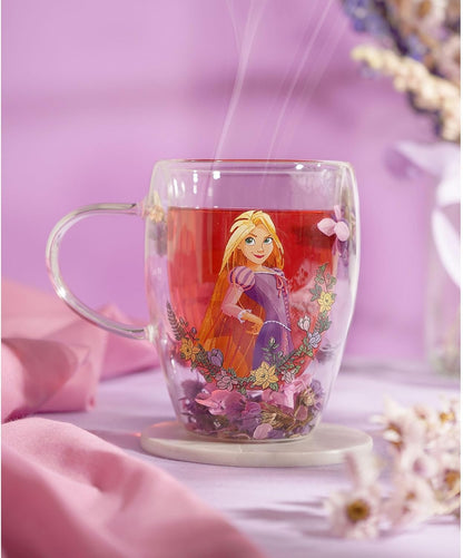 A clear glass mug with a printed illustration of Rapunzel from Tangled, filled with red tea and placed on a small white coaster against a soft pink background.