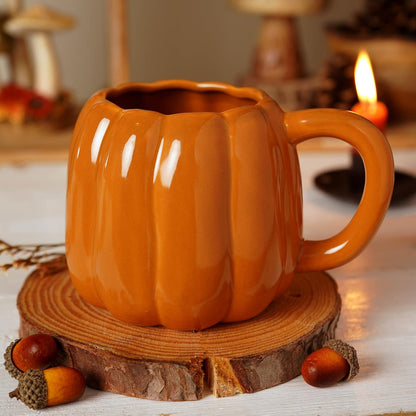 An orange pumpkin-shaped ceramic mug with a ribbed texture and a curved handle, resting on a rustic wood slice coaster with acorns and a warm candle in the background.