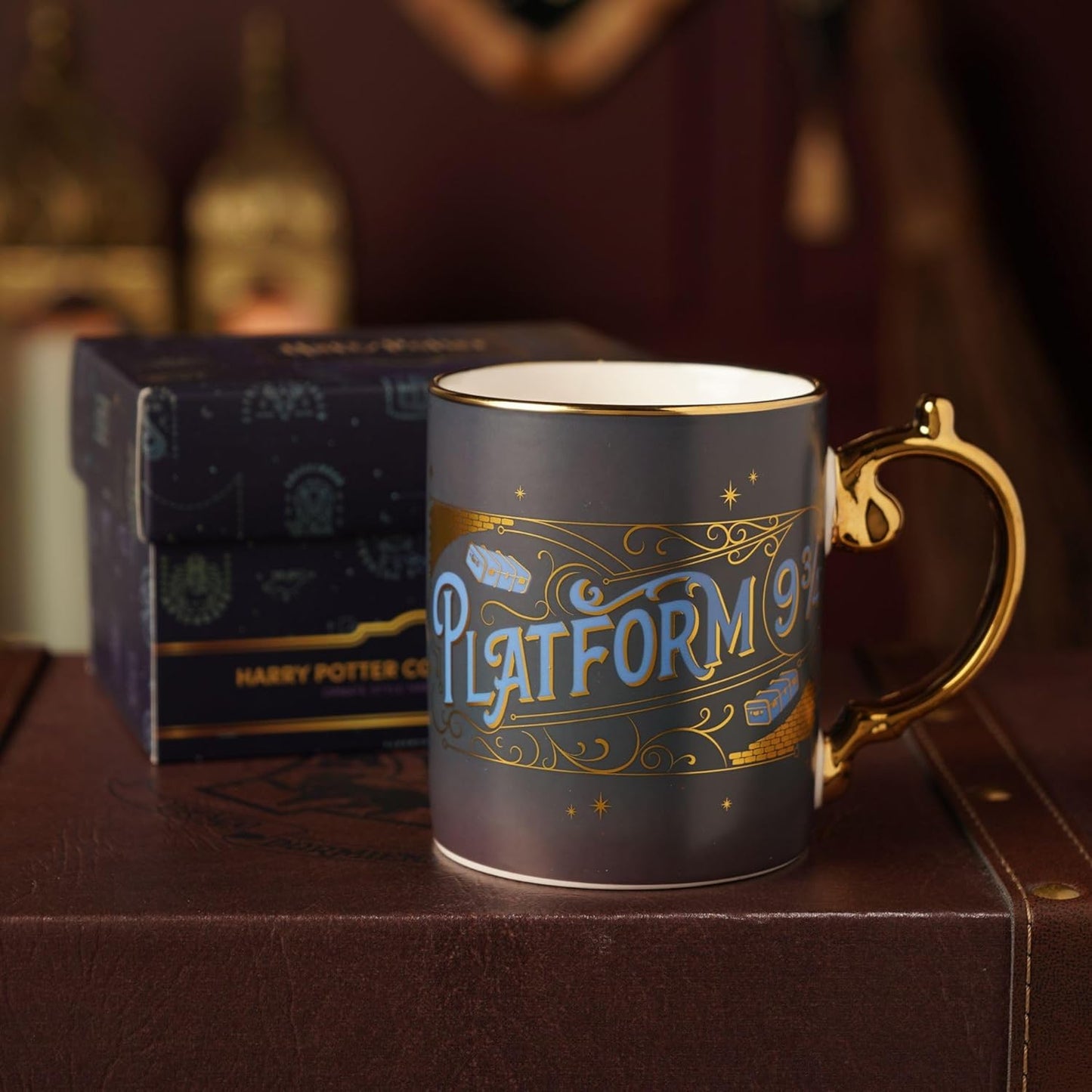 A Harry Potter themed ceramic mug featuring 'Platform 9 3/4' in blue and gold lettering, displayed next to its branded dark blue box on a brown leather surface.