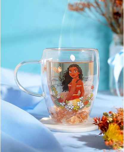 A clear double-walled glass mug with a handle, featuring an illustration of Moana from Moana Prime® on the front, set on a white coaster against a soft blue background with decorative dried flowers.