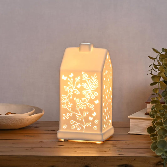 A glowing white ceramic house-shaped night light with floral cutout patterns, placed on a wooden table with a neutral background and decorative plants.