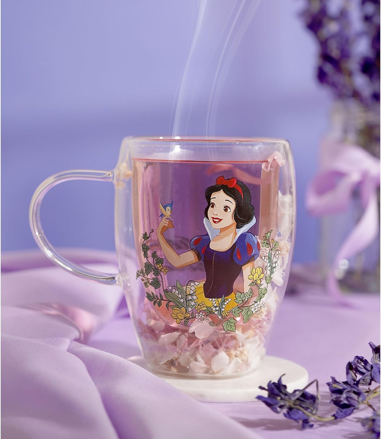A clear glass mug with a handle, featuring an illustration of Snow White on the front, filled with pink tea and flower petals, set against a soft lavender background with decorative flowers.