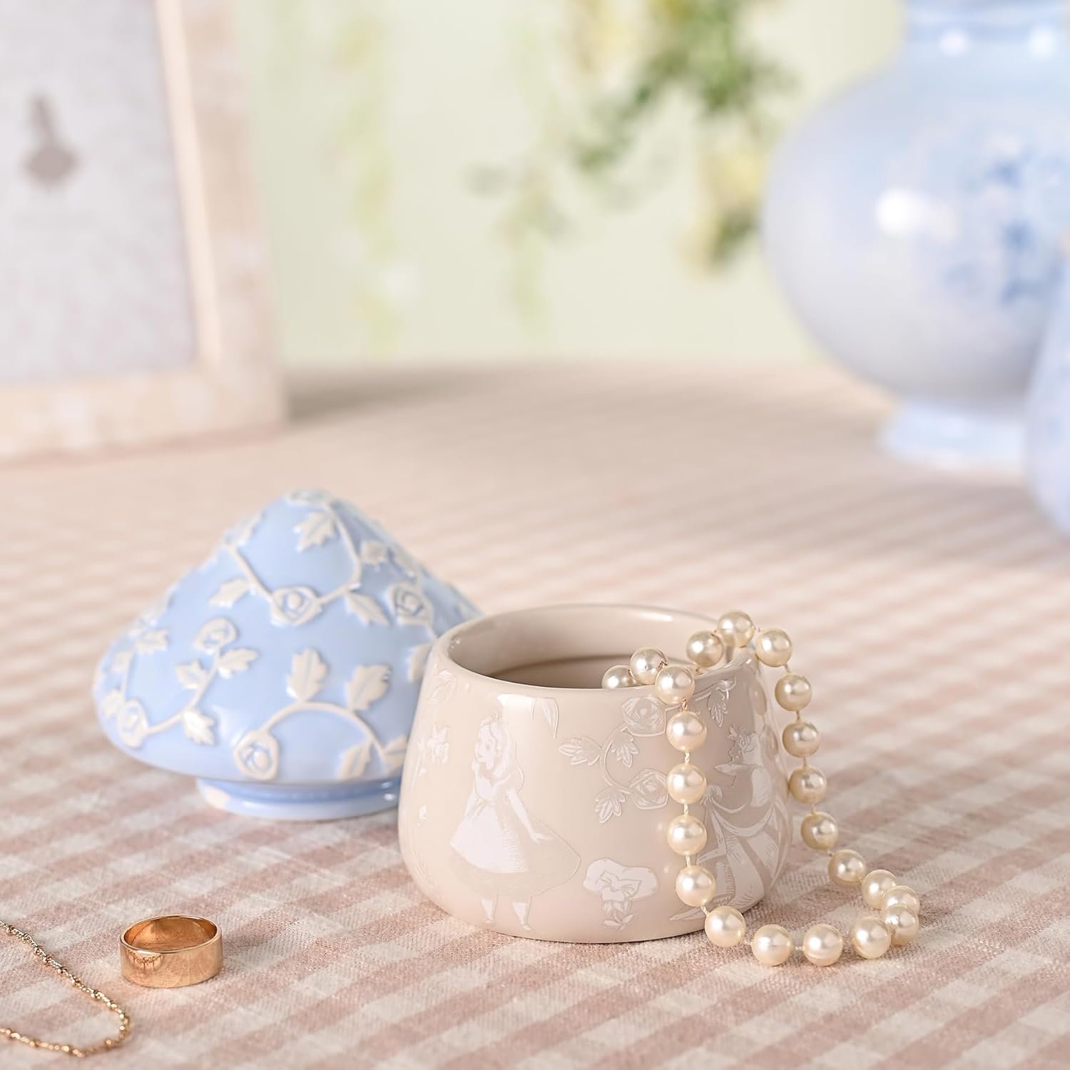 Ceramic container with floral design, pearl necklace, and gold ring on a textured surface.