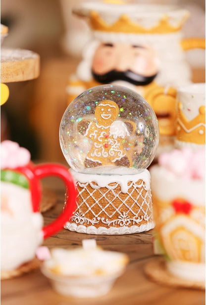 A gingerbread-themed snow globe with a decorated gingerbread figure inside, set on a patterned base. The scene is decorated with other gingerbread figurines and cups in a warm, festive setting.
