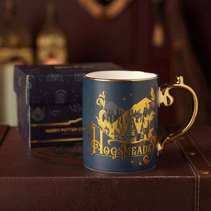 A navy blue ceramic mug with a gold Hogsmeade castle illustration and gold handle, shown in front of its matching branded box on a dark brown surface.