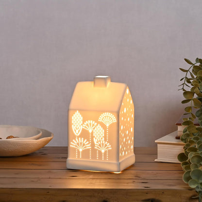 A small house-shaped ceramic night light with a warm white glow, featuring laser-cut fan and tree patterns on the front, displayed on a wooden tabletop with decorative props.