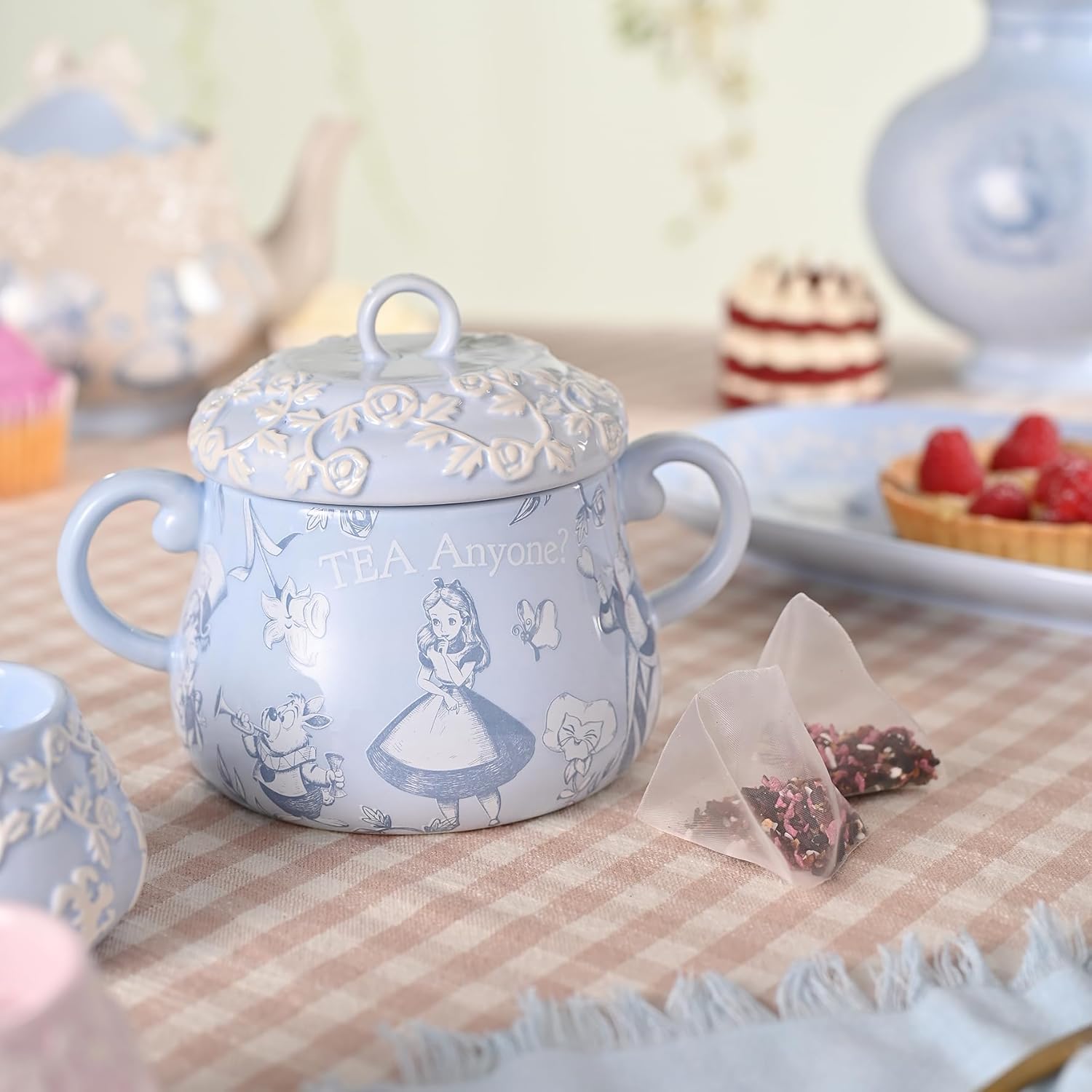 Decorative teapot with character design on a checkered tablecloth with tea bags and strawberries.