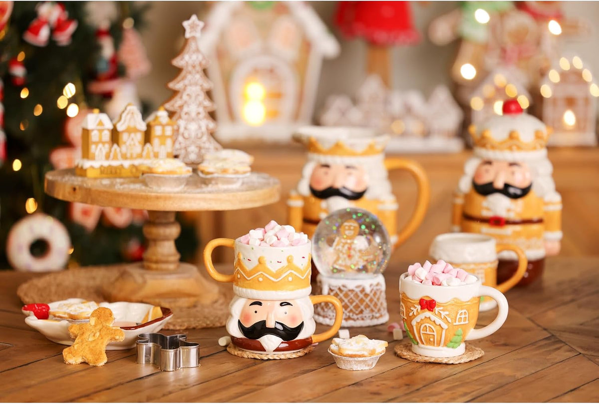A festive Christmas-themed ceramic mug and teapot set arranged on a wooden table, decorated with gingerbread houses, cookies, a snow globe, and warm holiday lights in the background.