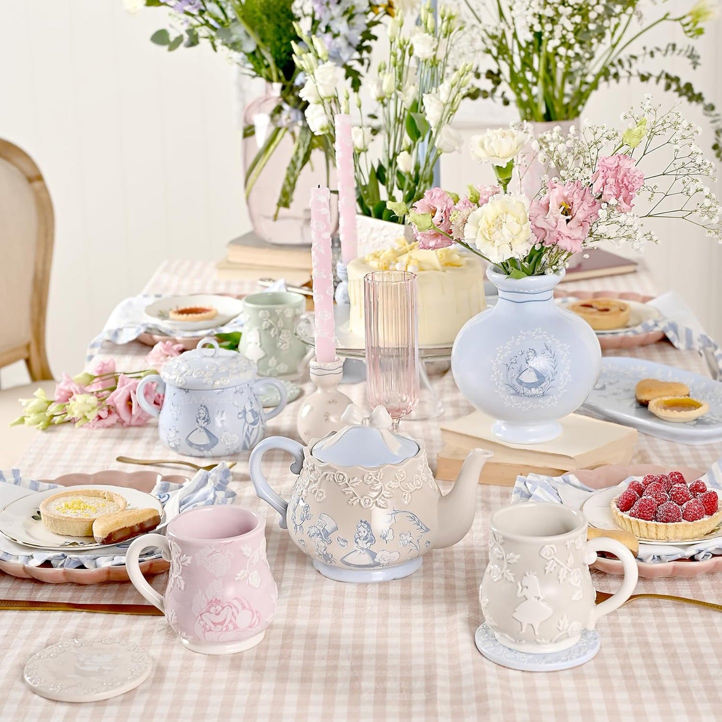 Pastel-colored table setting with teapots, mugs, and floral arrangements on a checkered tablecloth.