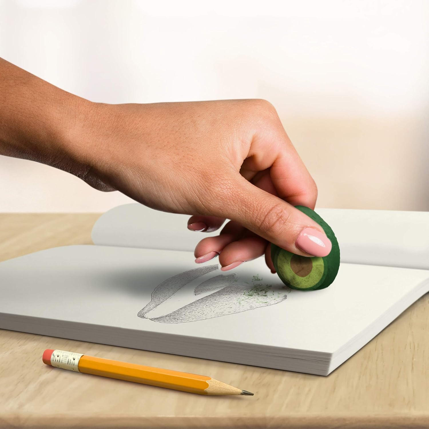 Hand holding a small avocado-shaped object over an open book with a pencil on a wooden surface.